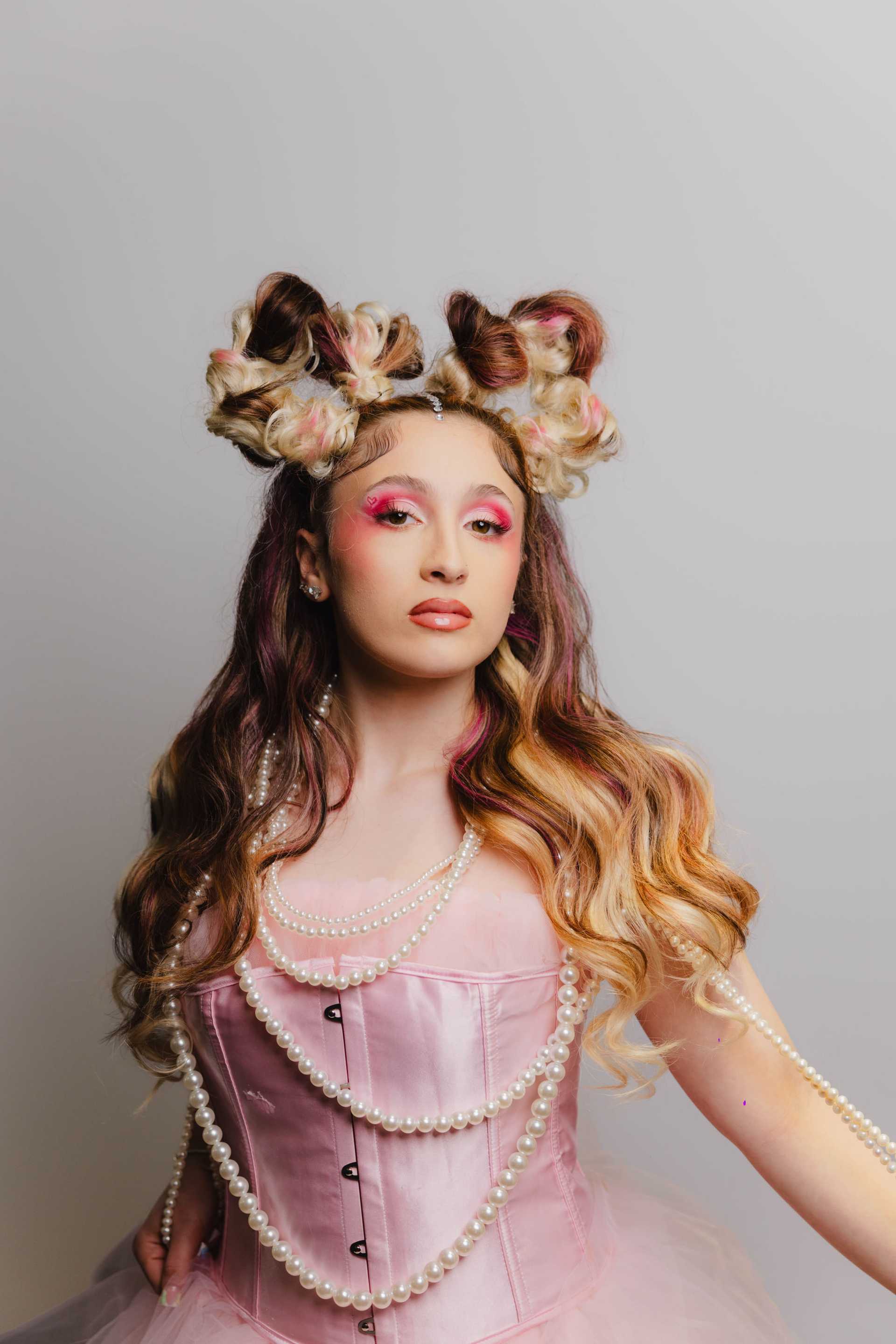 Young woman in elaborate hairstyle and pink dress with pearls, against a gray background.