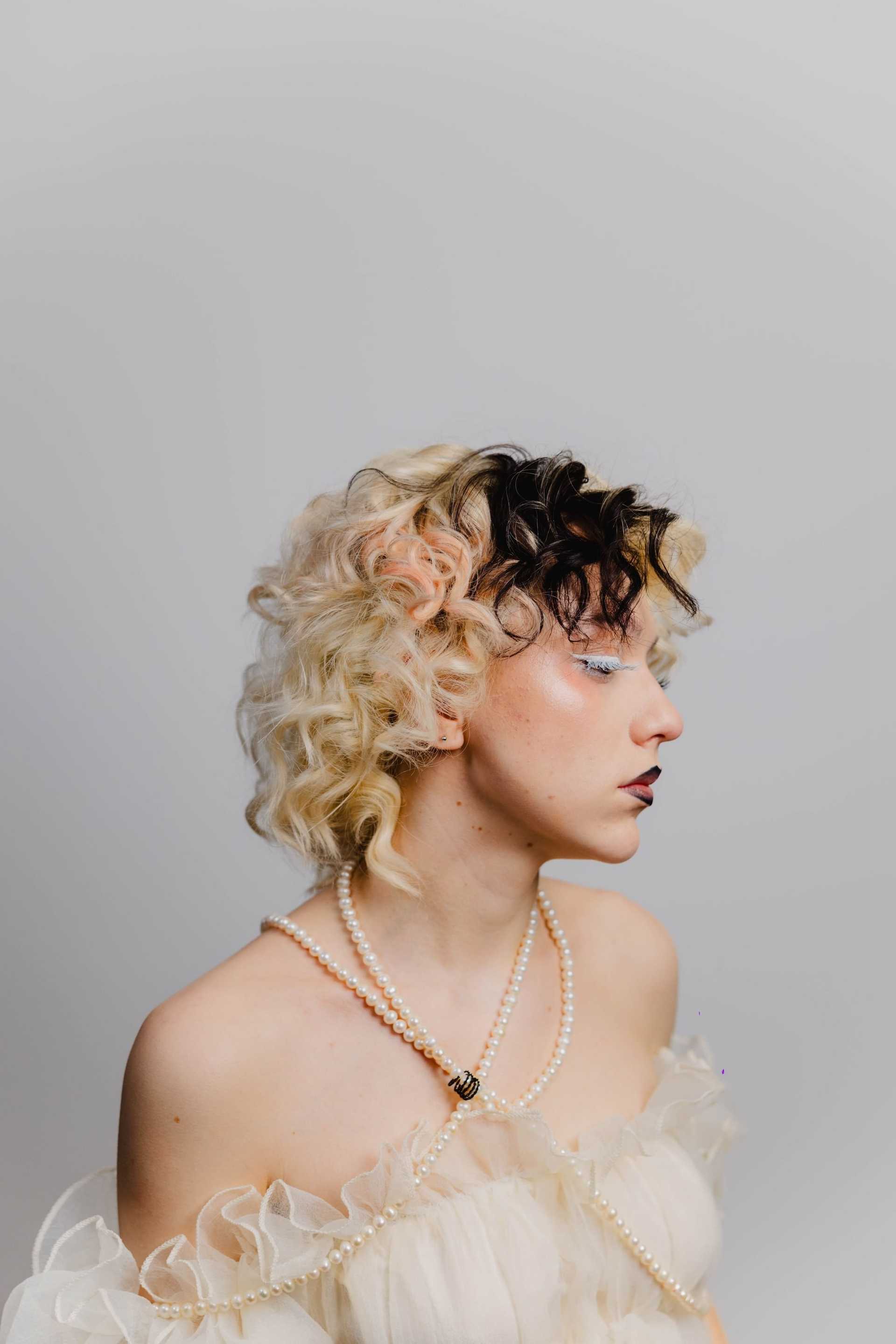 Model with curly, two-toned hair and pearl necklace against a neutral background.