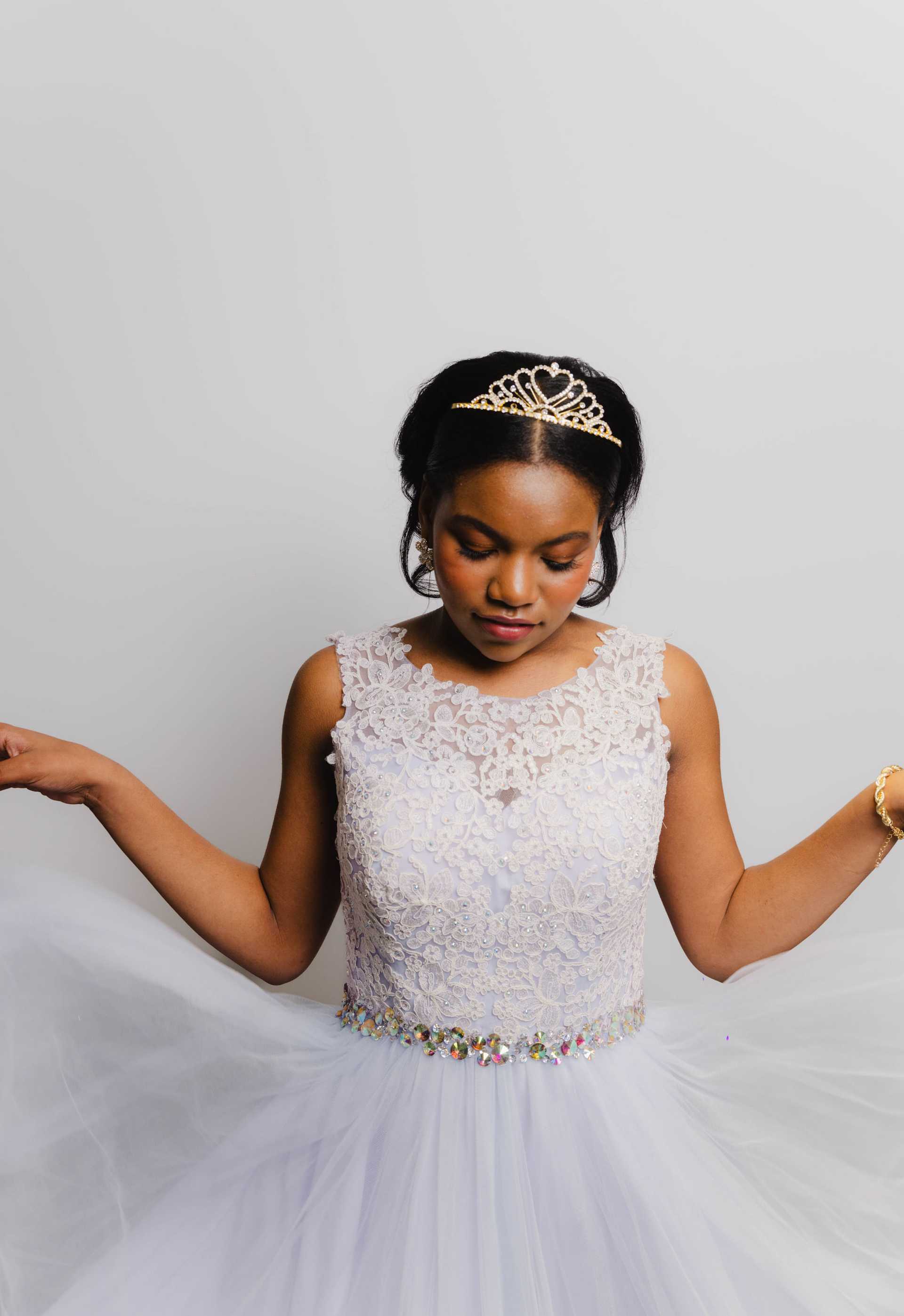 Young woman in white lace dress and tiara, posing gracefully against a plain background.
