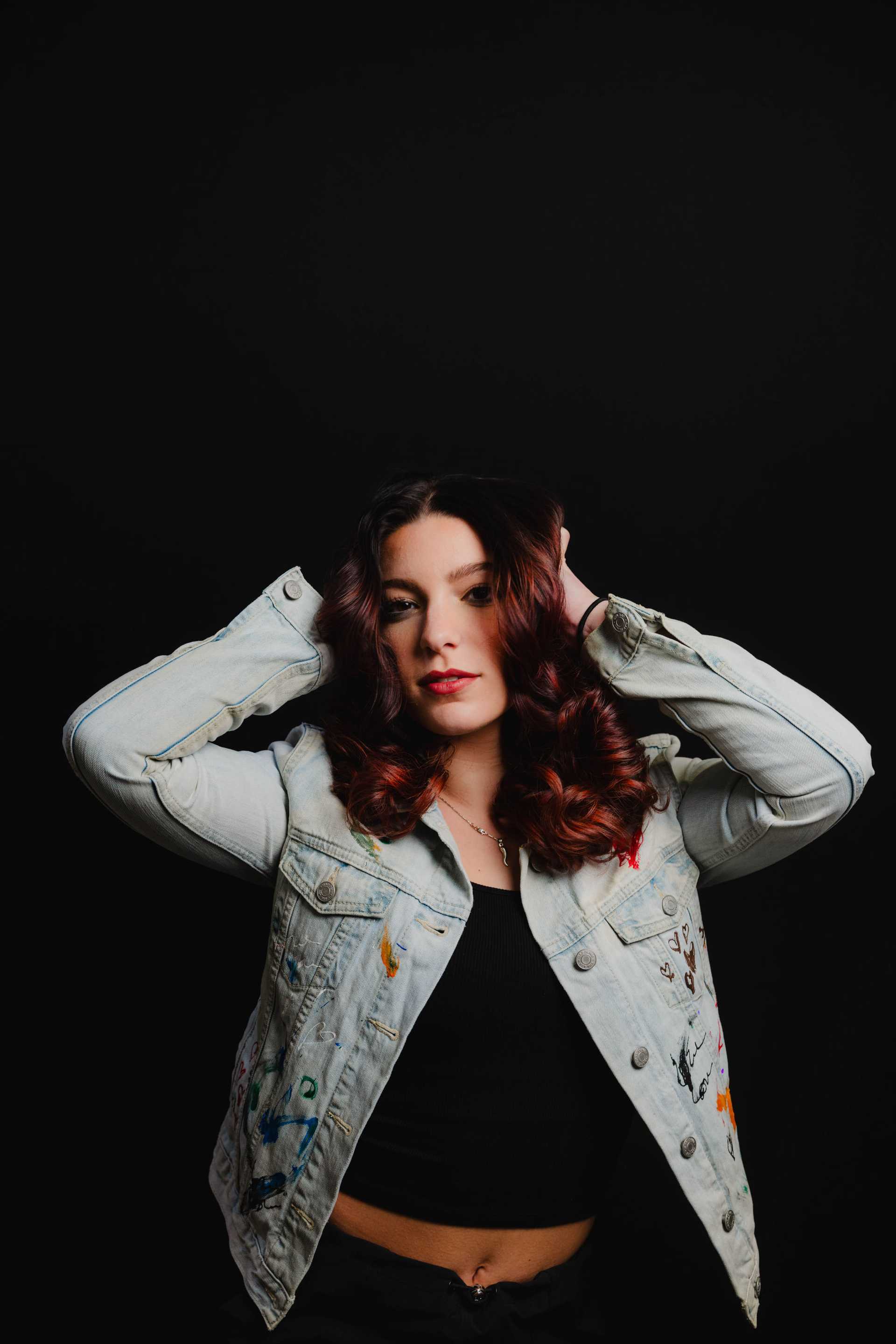 Woman with wavy red hair poses in a painted denim jacket against a black background.