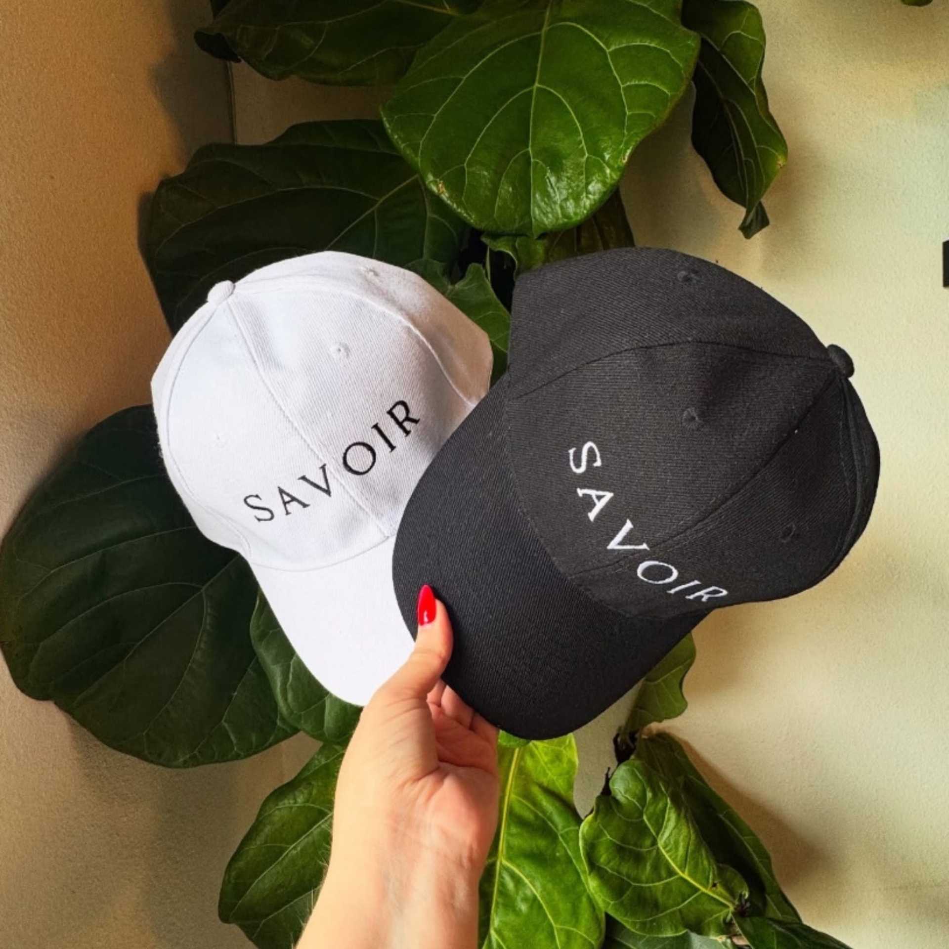 Two caps, one white and one black, with "Savoir" text, held in front of large green leaves.
