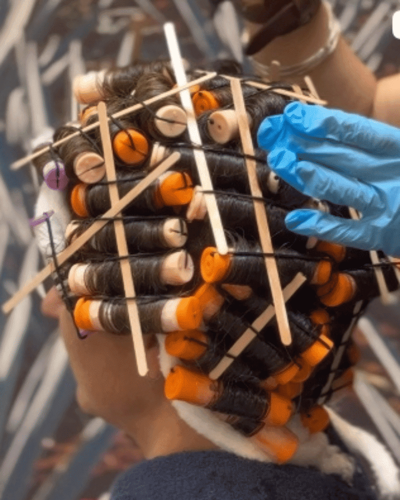 Hair in curlers with wooden sticks, person wearing a towel and blue gloves adjusting.