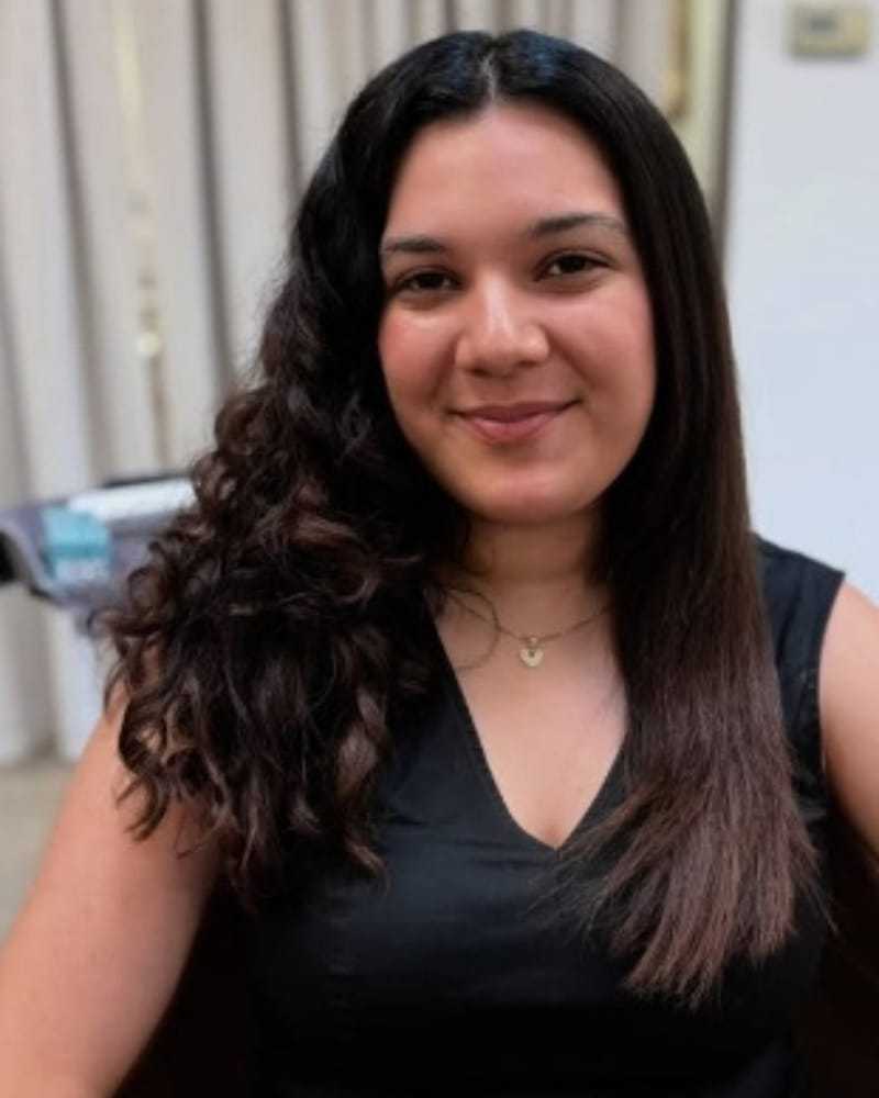 Smiling woman with half curly, half straight hair in a black top, indoors.