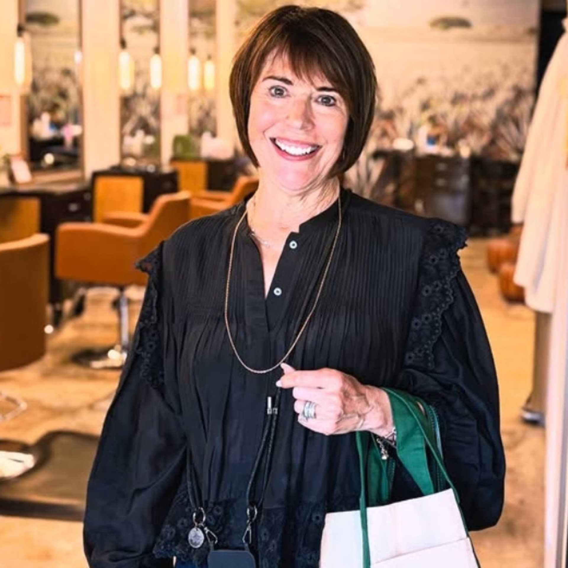 Woman smiling in a salon, holding a shopping bag, wearing a black blouse.