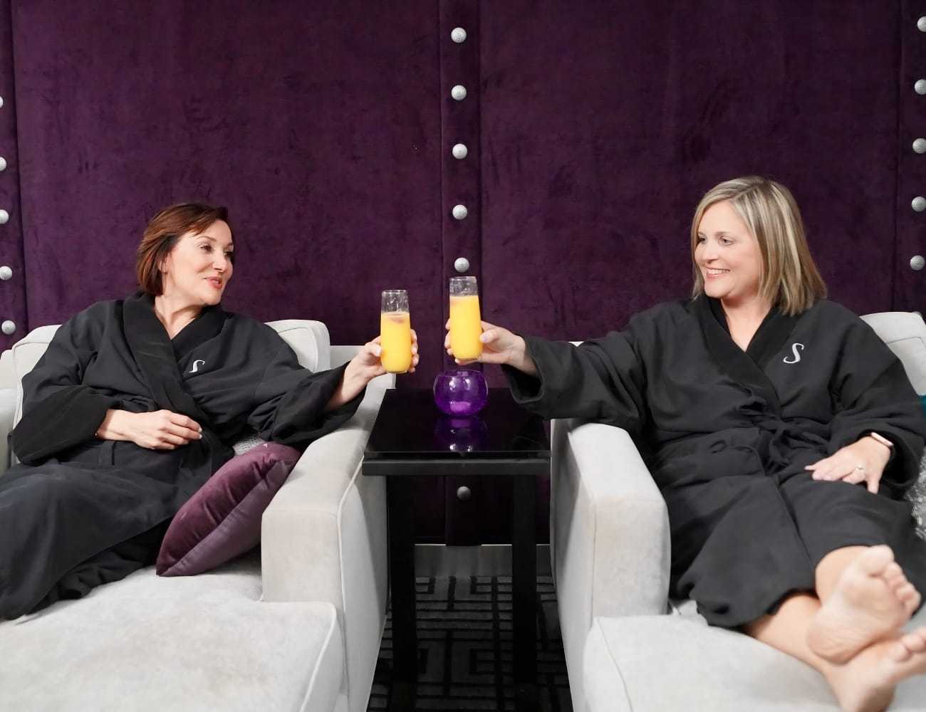 Two women in robes toasting with orange drinks, relaxing on lounge chairs.