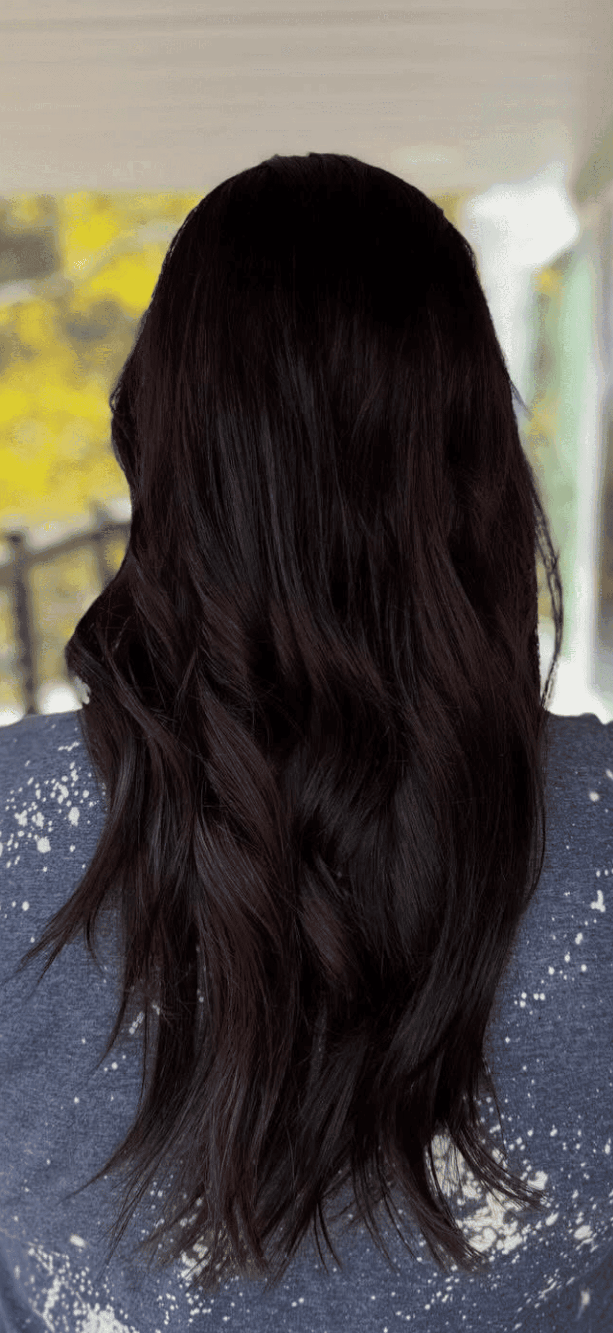 Back view of a person with long, wavy dark hair, wearing a blue speckled shirt.