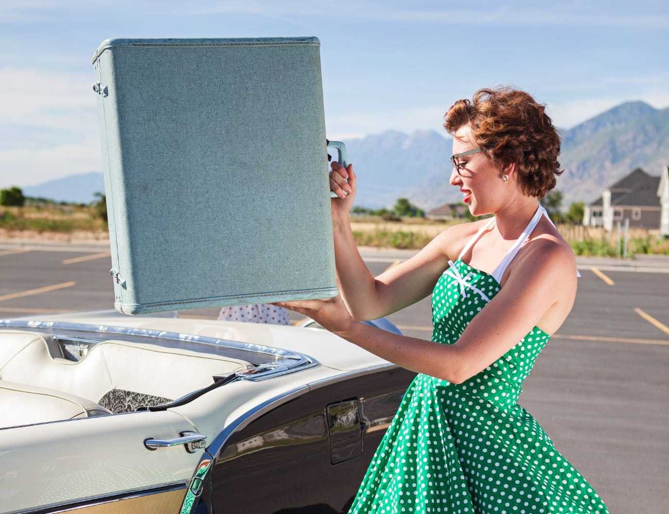 Woman in green dress holding a blue suitcase by a classic convertible car.