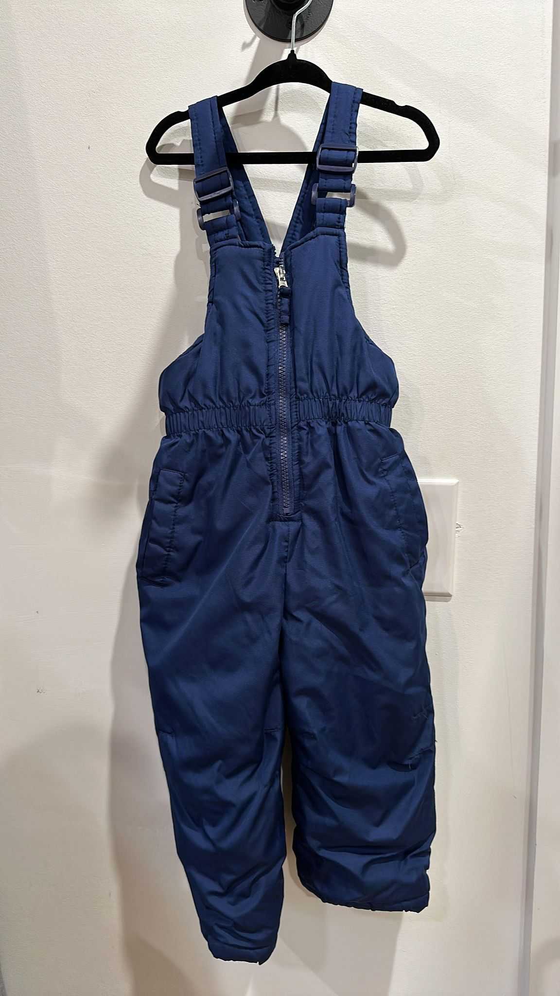 Blue snow overalls hanging on a black hanger against a white wall.