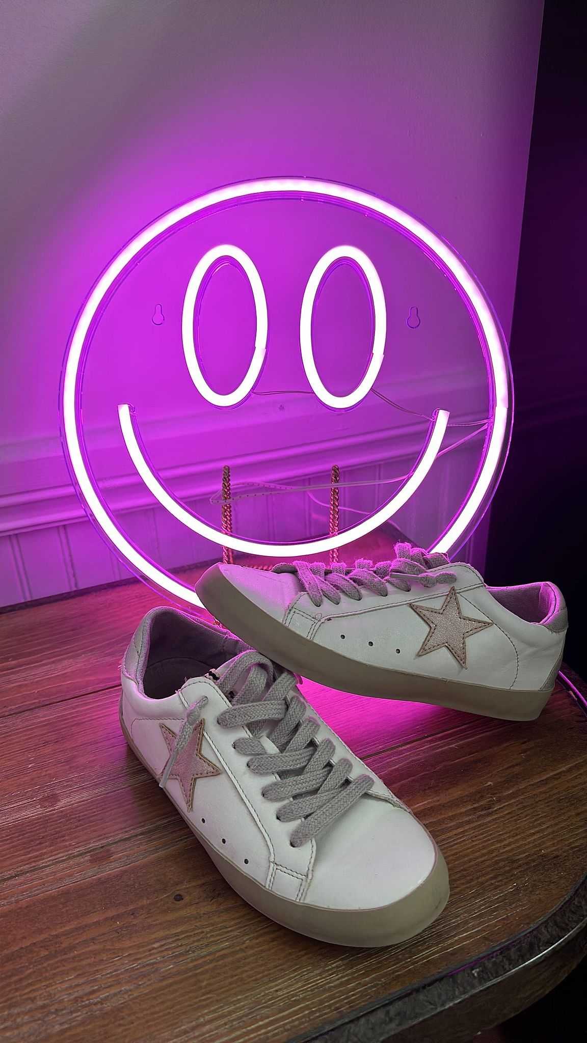 White sneakers on a wooden surface with a neon purple smiley face sign in the background.