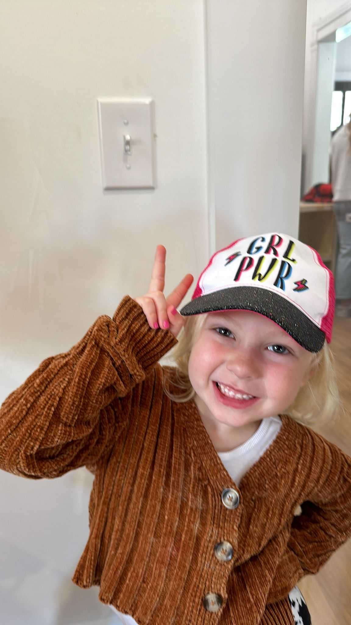 Child smiling, wearing a "GRL PWR" cap and a brown knit sweater, making a peace sign.