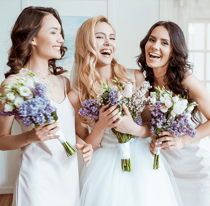 Three joyful bridesmaids holding bouquets, smiling and laughing together indoors.