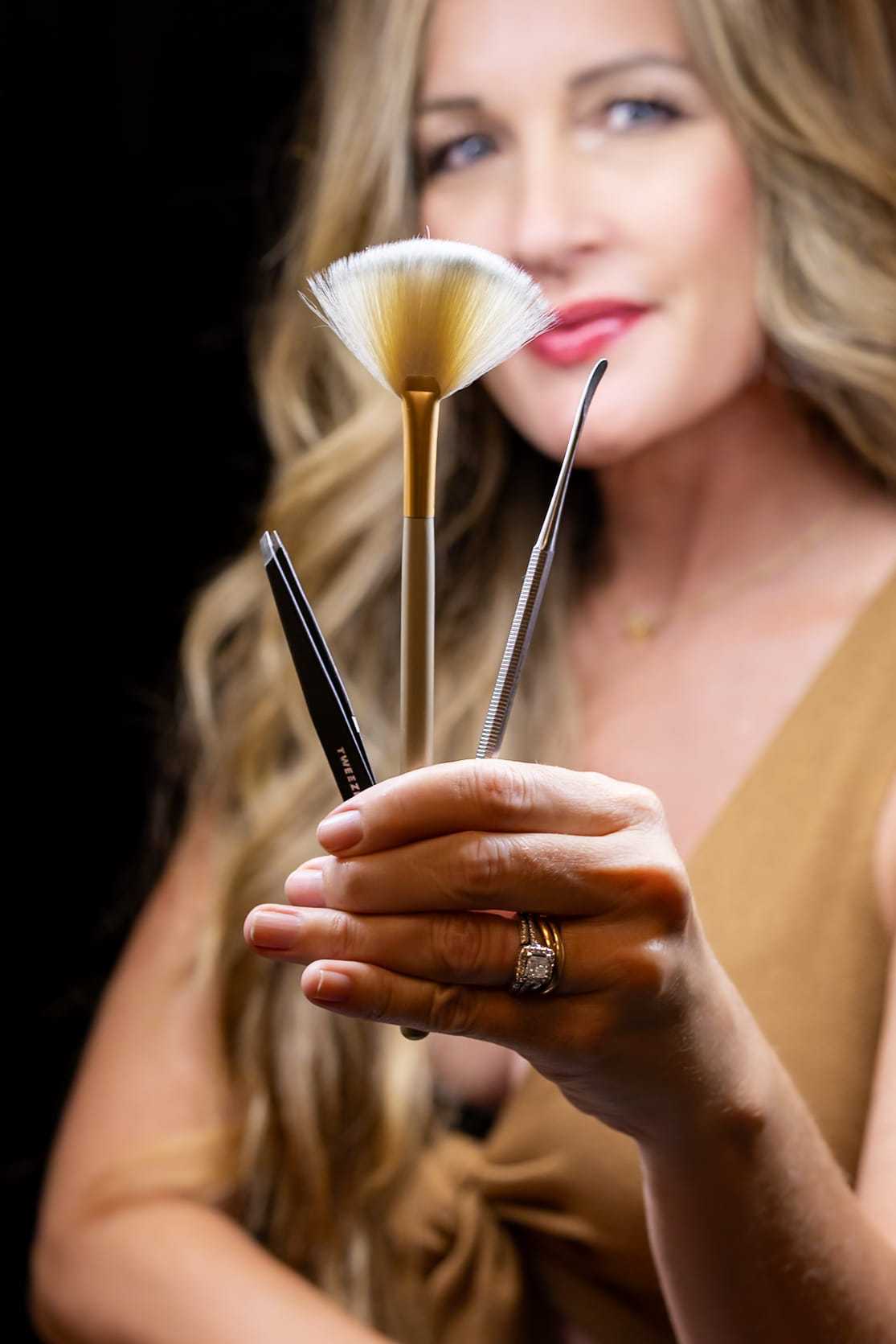 Woman holding beauty tools: fan brush, tweezer, and lash applicator.