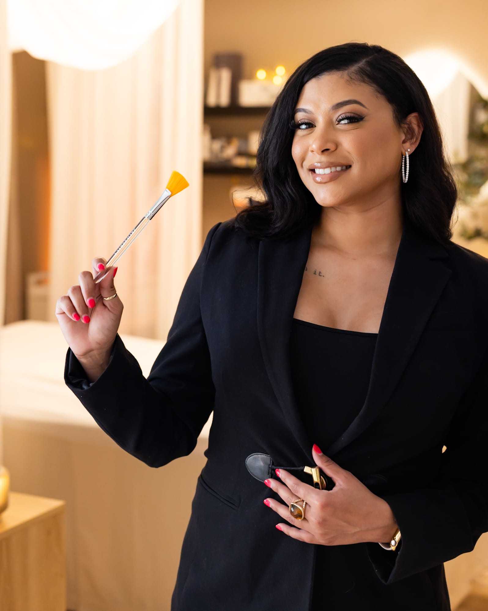 Woman in black blazer holding a small brush, smiling in a softly lit room.