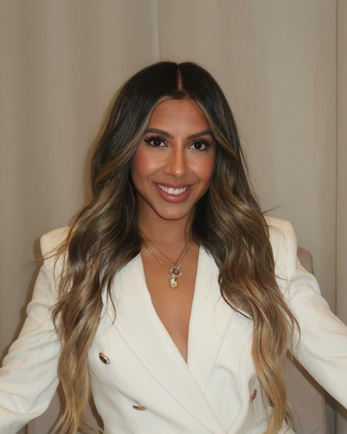 Young woman with long hair smiles, wearing a white blazer against a neutral background.