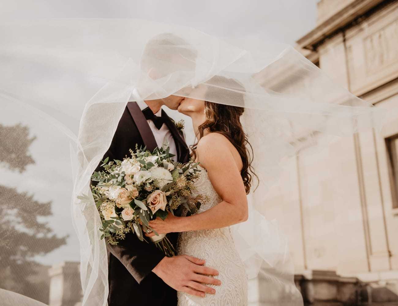Bride and groom kiss under veil, holding bouquet in front of elegant building.