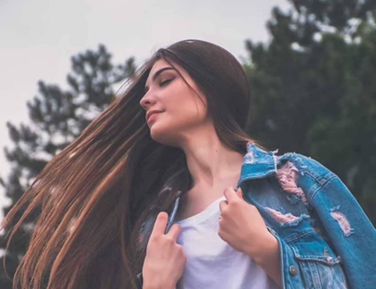 Young woman with long hair, eyes closed, wearing a denim jacket in a wooded area.