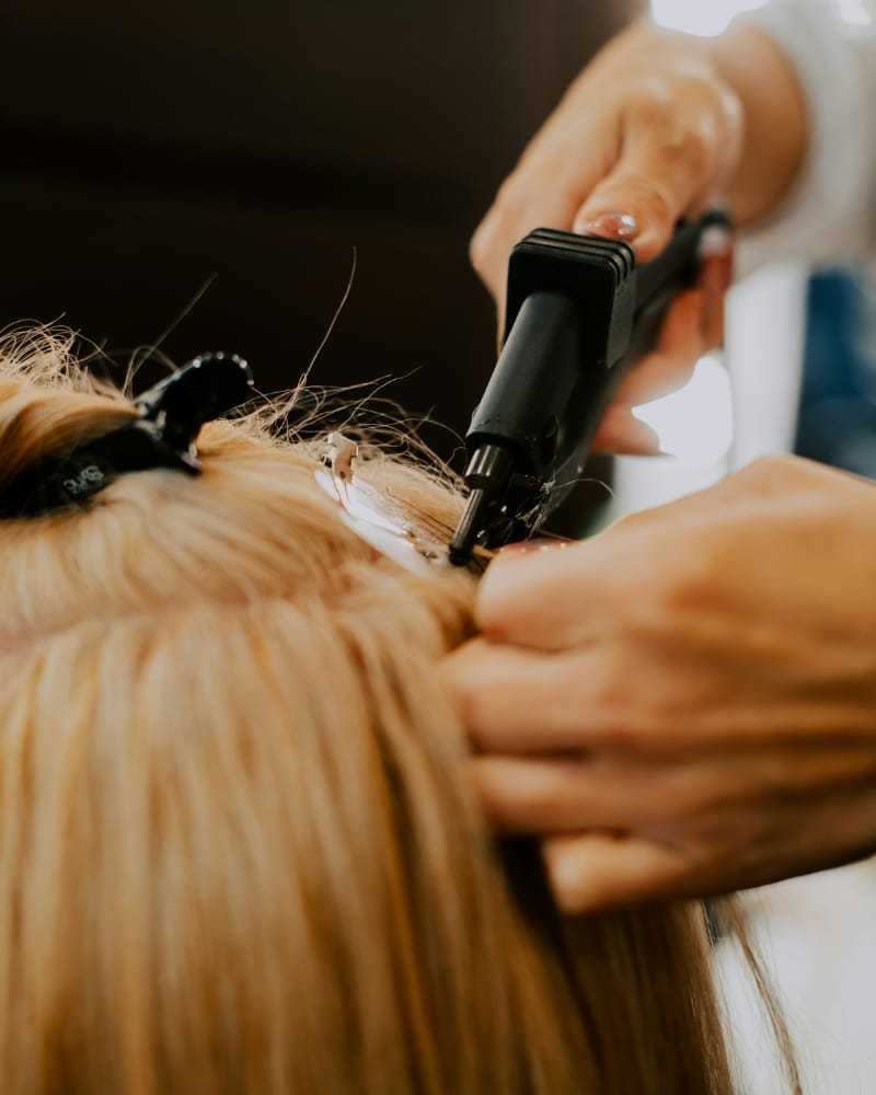Hairdresser applying extensions to blonde hair with heated tool.